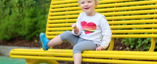 Child Enjoying A Snack On A Bright Yellow Park Bench In A Lush Green Setting.