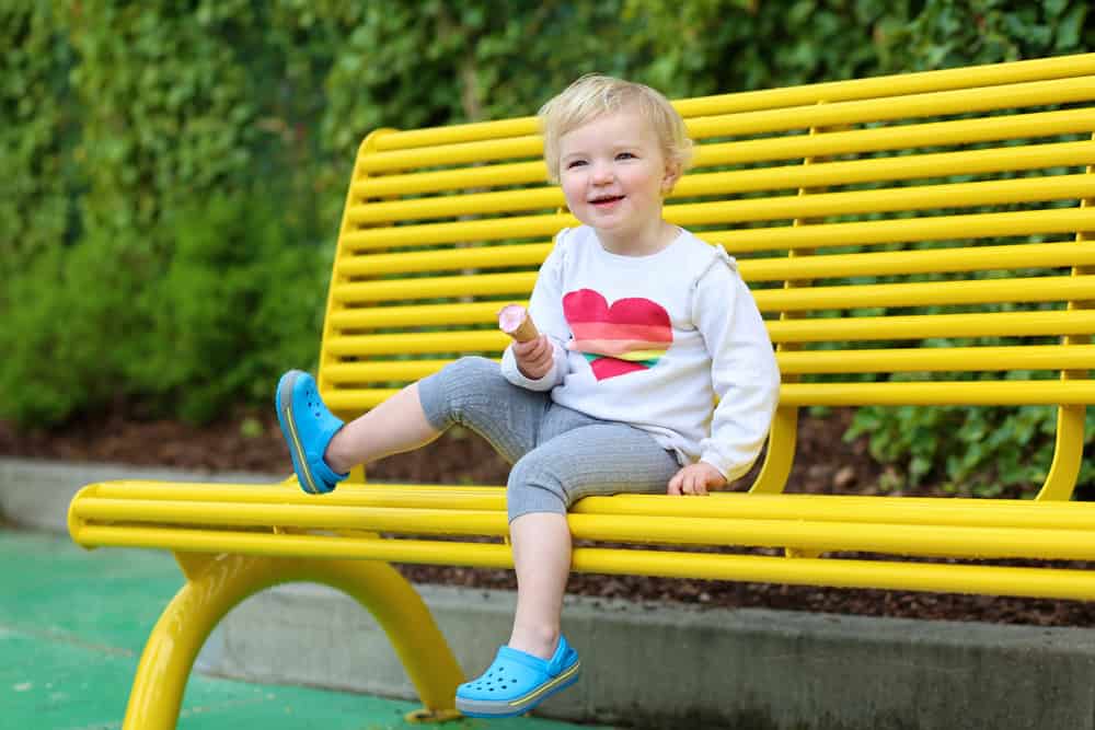 Child enjoying a snack on a bright yellow park bench in a lush green setting.