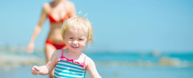 Smiling Young Girl In A Striped Swimsuit Playing On The Beach With A Woman In The Background.