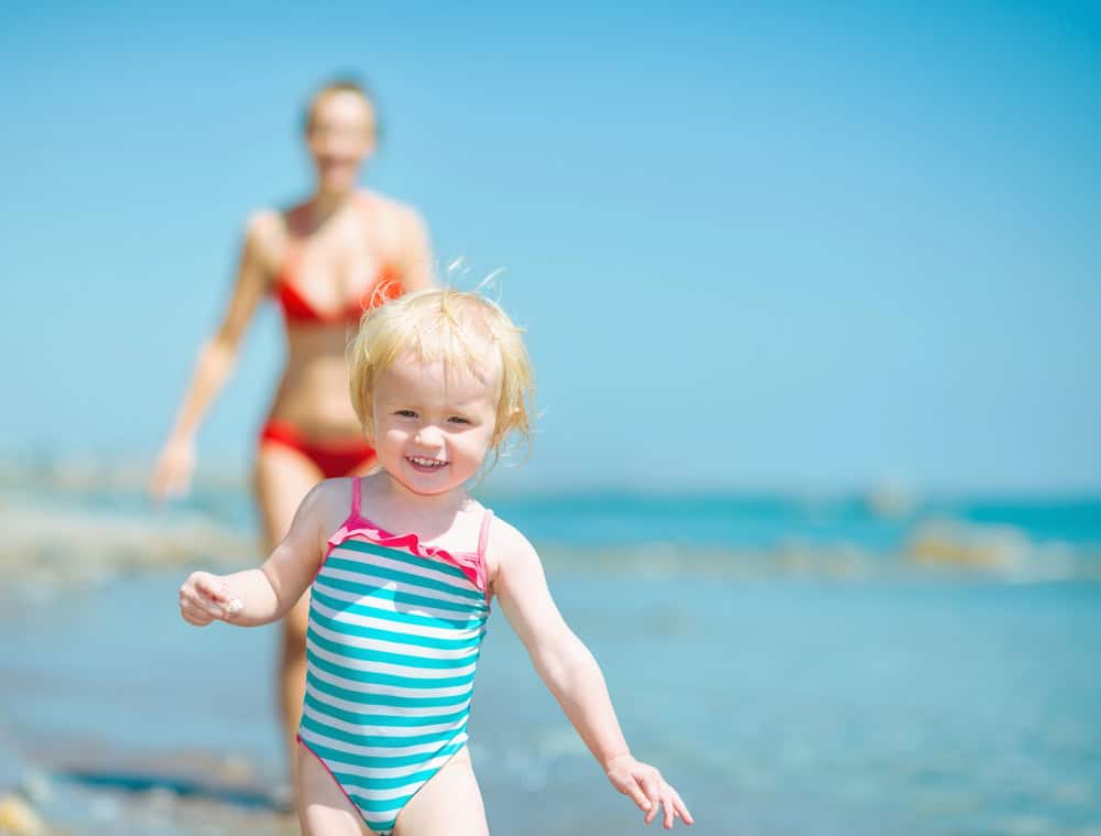 Smiling young girl in a striped swimsuit playing on the beach with a woman in the background.
