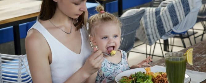 A Woman Feeding A Young Girl At A Restaurant Table With Healthy Food And A Green Smoothie.