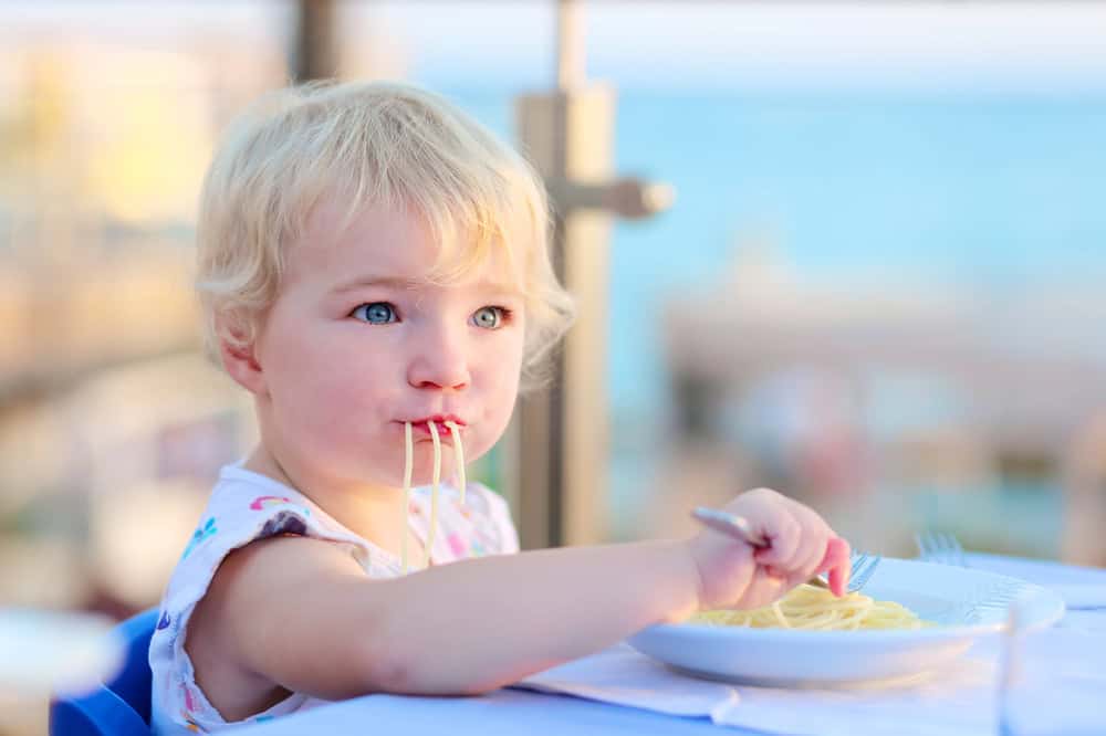 Young child eating spaghetti outdoors with ocean view in background.