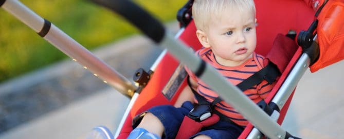 Baby In Red Stroller Outdoors, Child With Blonde Hair And Striped Shirt, Exploring On A Walk.