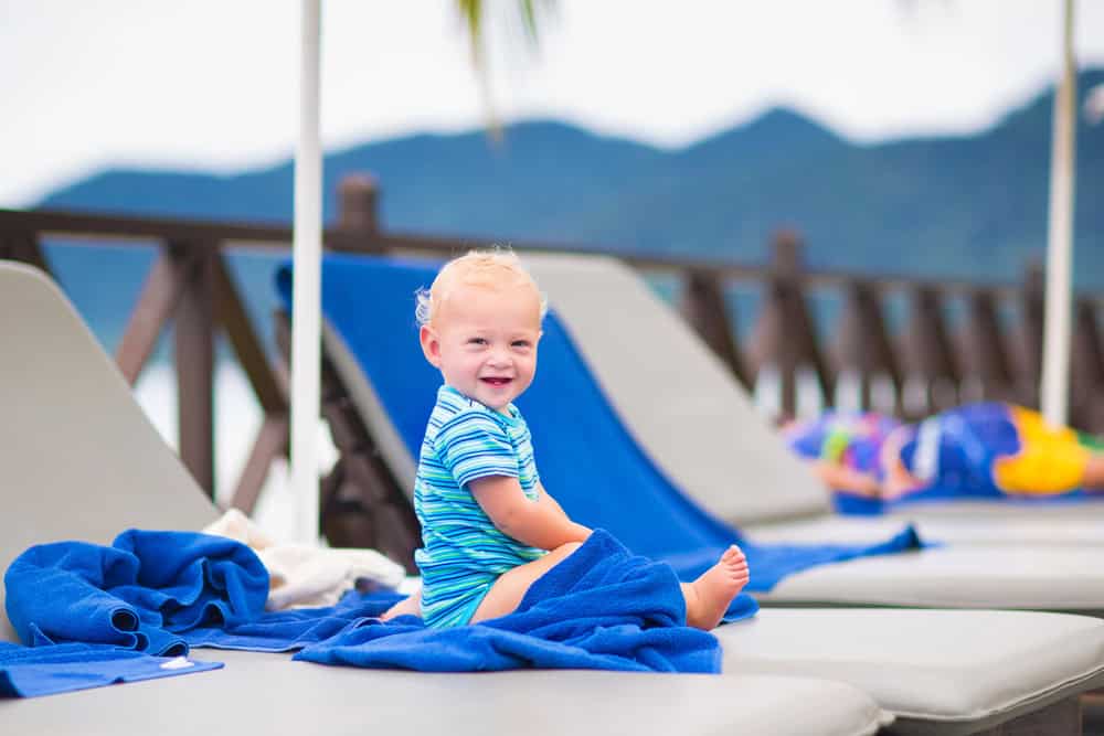 Smiling toddler sitting on poolside lounge chair with mountain view.