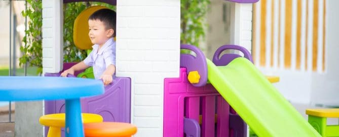 Child Playing In Colorful Plastic Playhouse With Slide And Outdoor Furniture.