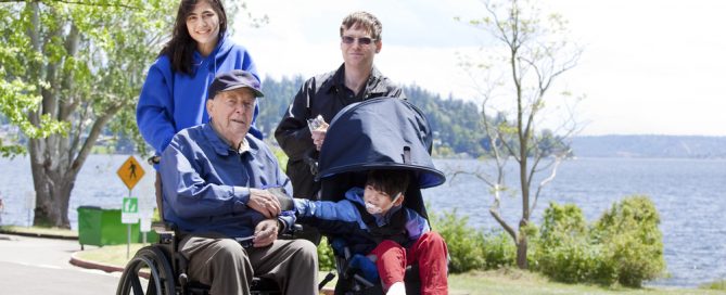 Family Enjoying Outdoor Walk With Senior In Wheelchair By Water.