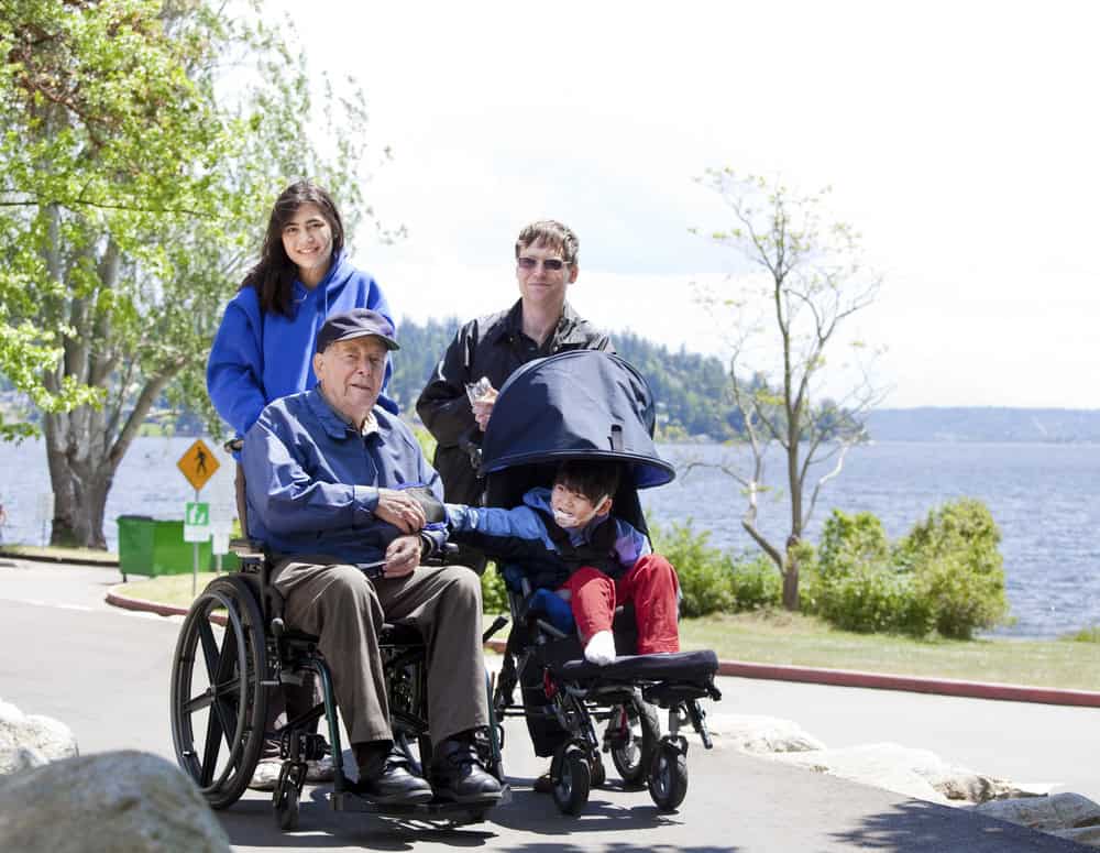 Family enjoying outdoor walk with senior in wheelchair by water.