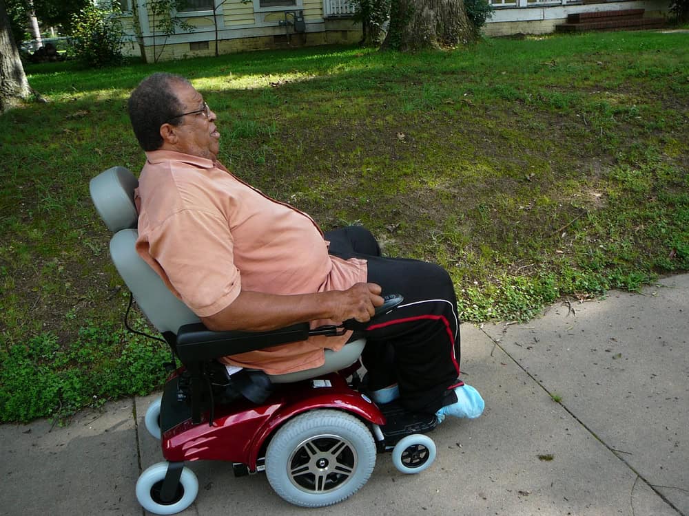 Senior man in a motorized wheelchair outdoors on a sidewalk.