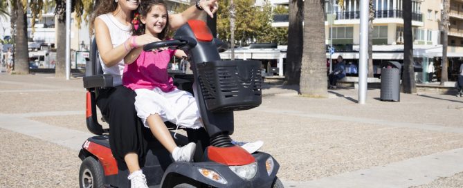 Mother And Daughter Riding A Mobility Scooter In A Park.