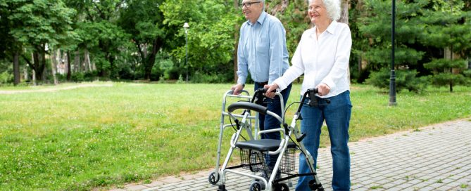 Elderly Couple Walking Outdoors With A Walker In A Park Setting.