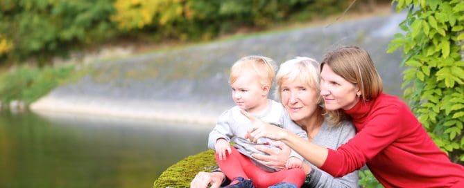 Elderly Woman With Young Girl And Caregiver By A River In A Park.