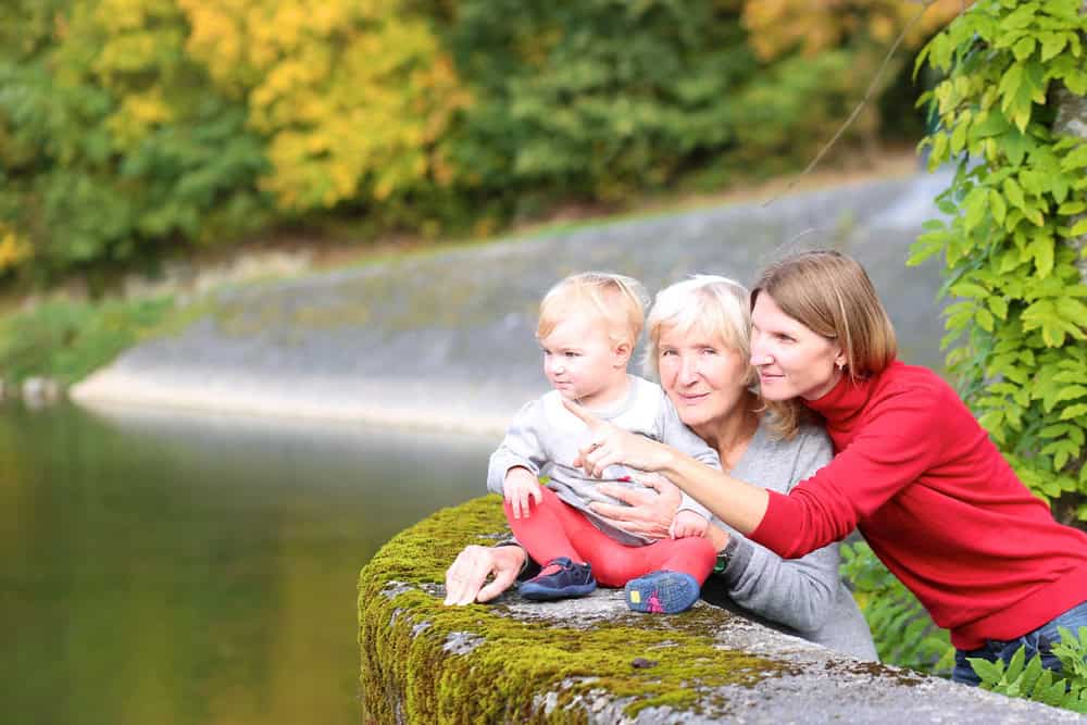 Elderly woman with young girl and caregiver by a river in a park.