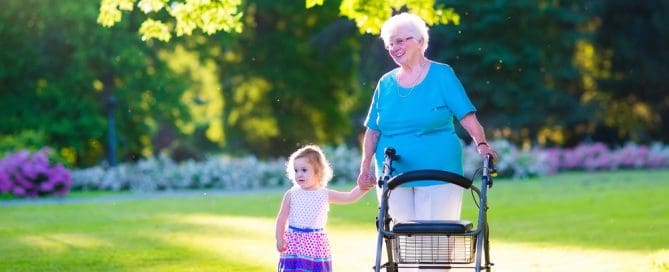 Elderly Woman With Walker Walking With Young Girl In Park On Sunny Day.
