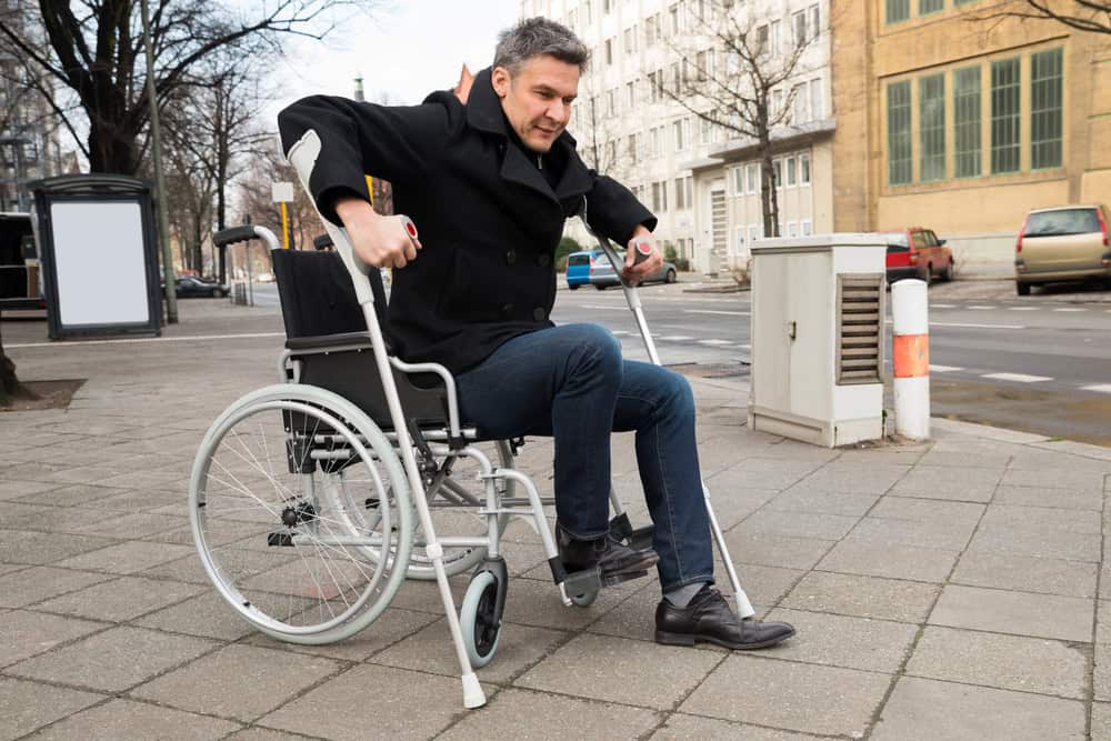Man using crutches to transfer from wheelchair on city sidewalk.