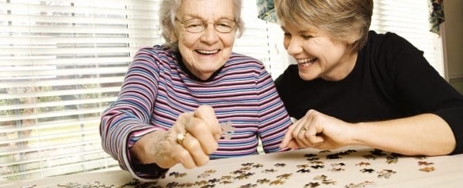 Elderly Woman And Caregiver Solving A Puzzle Together At Home.