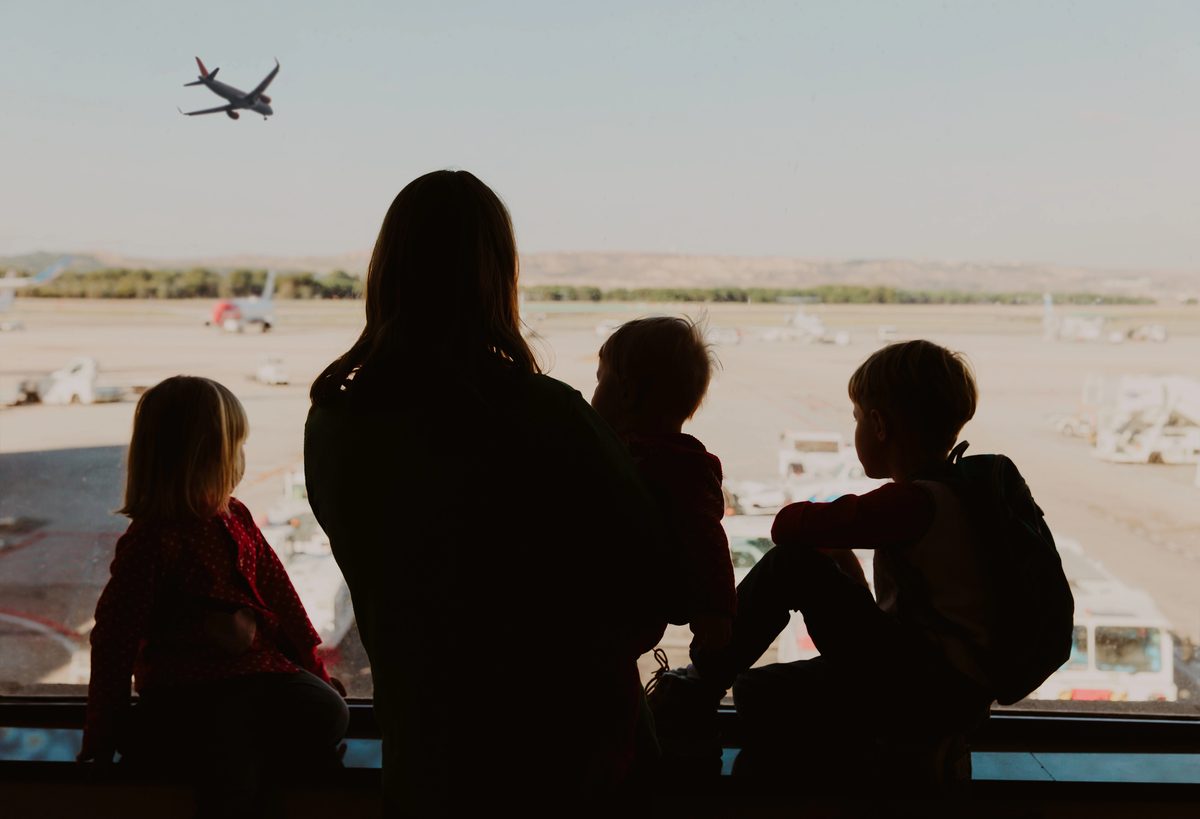 Mother And Three Young Children Watching Planes From An Airport Window On A Travel Day
