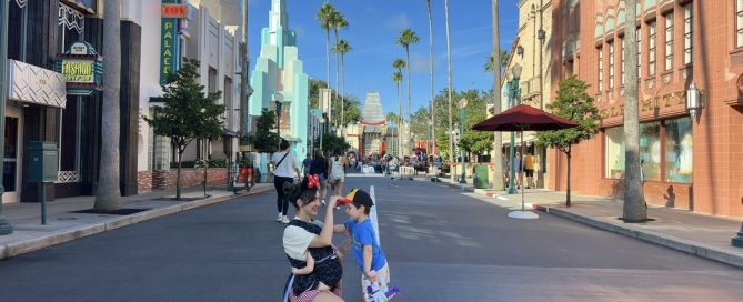 A Mom Carrying Her Baby Kneels Beside Her Toddler On Hollywood Boulevard At Disney'S Hollywood Studios During A Disney World Trip With A Baby And Toddler.