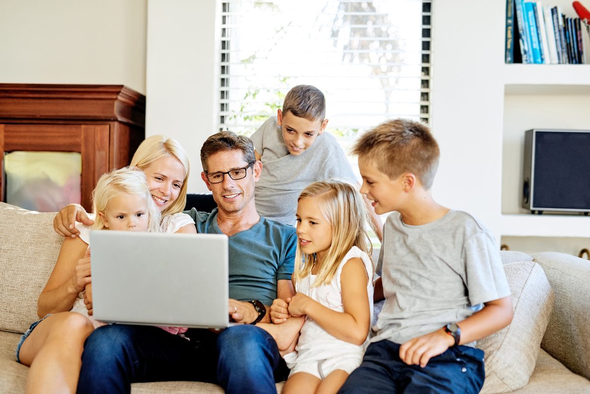 Parents And Four Kids On A Couch Looking At A Laptop Together While Planning A Trip